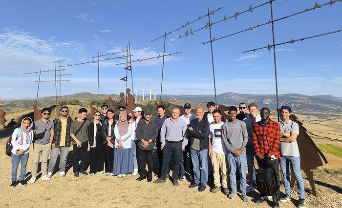 Group photo beside a metal sculpture installation, wind turbines in the distance, 2025